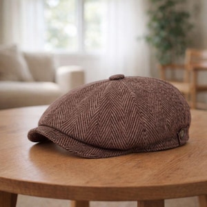 Puede incluir: Gorra estilo newsboy marrón con estampado de espiga, visera pequeña y botón en la parte superior. La gorra está hecha de tela texturizada y se encuentra sobre una mesa de madera. Tiene un estilo clásico y vintage.