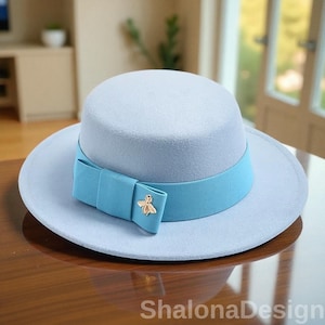 Può includere: Un cappello azzurro con tesa larga e un nastro blu abbinato. Il nastro è annodato a fiocco e presenta un ciondolo a forma di ape dorata. Il cappello è su una superficie di legno. Il testo "ShalonaDesign" è in basso.