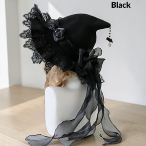 May include: A black witch hat featuring a wide brim adorned with intricate black lace trim. The hat is decorated with a black satin ribbon bow and a sheer organza flower. A dangling charm featuring a small black bat and a star hangs from the tip.
