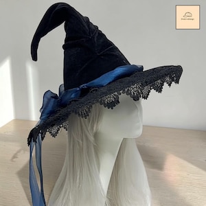 Puede incluir: Un sombrero puntiagudo de terciopelo negro con un ala ancha que presenta un intrincado borde de encaje negro. Una cinta transparente de color azul intenso rodea la base de la copa, atada en un lazo lateral con extremos largos que cuelgan.