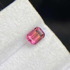 May include: A rectangular-cut, pink gemstone with a gradient of colors from deep pink to a lighter peach hue. The gemstone is set against a white, textured background, with a dark background.