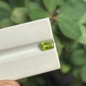 Peut inclure: Une pierre précieuse rectangulaire vert clair, possiblement de la péridot, est présentée sur une surface blanche. La pierre est tenue devant un fond de feuillage vert flou. La coupe et la couleur de la pierre sont clairement visibles.