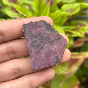 Könnte beinhalten: Ein flaches, unregelmäßig geformtes Stück roher Rubin mit einem tiefen Magenta- und Violettton. Die Oberfläche weist eine raue, unpolierte Textur mit sichtbaren dunklen Einschlüssen und natürlichen Mineraladern auf der Steinvorderseite auf.