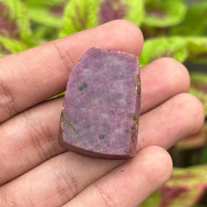 May include: A raw, unpolished ruby gemstone with a flat, irregular shape and a deep magenta-pink hue. The surface displays a natural, textured crystalline structure with visible inclusions and subtle variations in color throughout the stone.