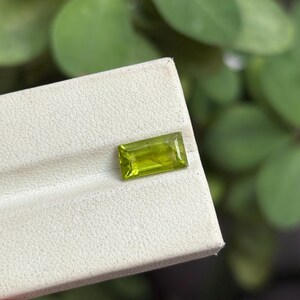 Può includere: Una gemma rettangolare verde chiaro con un aspetto luminoso e traslucido. La gemma è posta su una superficie bianca, con uno sfondo verde sfocato. La gemma è probabilmente un peridoto, una scelta popolare per i gioielli.