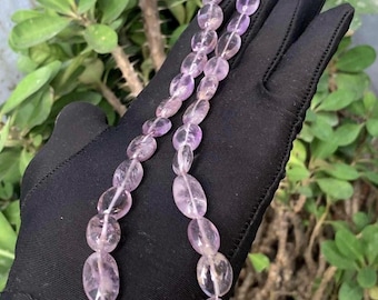 May include: A necklace of oval-shaped amethyst beads. The beads are a pale purple color and are strung together on a clear string. The necklace is held against a black background, with green foliage in the background.