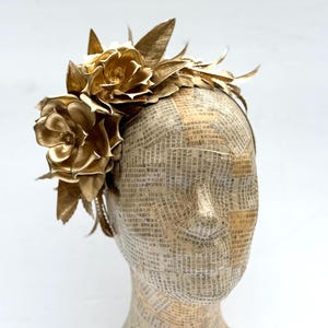May include: A golden headpiece featuring large, metallic-looking flowers and leaves. The headpiece is attached to a headband. The mannequin head is covered in paper with printed text.