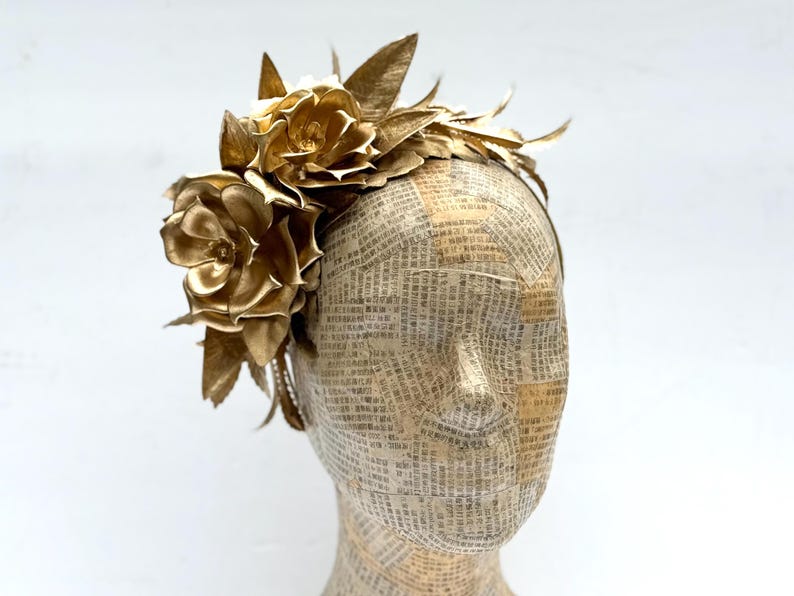 May include: A golden headpiece featuring large, metallic-looking flowers and leaves. The headpiece is attached to a headband. The mannequin head is covered in paper with printed text.