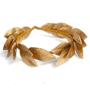 May include: A gold laurel wreath made of fabric leaves. The wreath is a circle and is lying flat on a white surface.