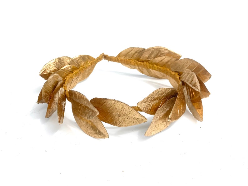 May include: A gold laurel wreath made of fabric leaves. The wreath is a circle and is lying flat on a white surface.