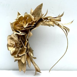 May include: A golden floral headpiece featuring two large, detailed flowers and delicate leaf accents. The headpiece has a thin, flexible band for comfortable wear. The overall design is elegant and suitable for special occasions.