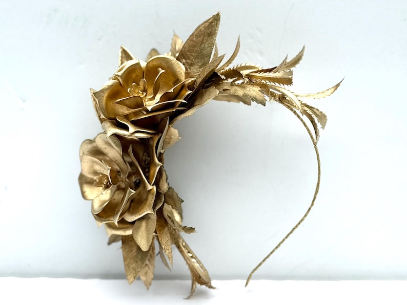 May include: A golden floral headpiece featuring two large, detailed flowers and delicate leaf accents. The headpiece has a thin, flexible band for comfortable wear. The overall design is elegant and suitable for special occasions.