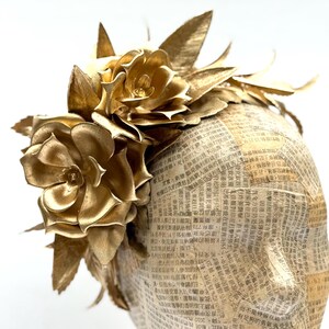 May include: A golden headpiece featuring large, metallic gold flowers and leaves. The base is covered in newspaper print. The floral arrangement is clustered on top, creating a striking, elegant design.