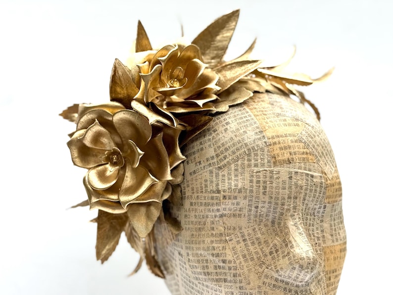 May include: A golden headpiece featuring large, metallic gold flowers and leaves. The base is covered in newspaper print. The floral arrangement is clustered on top, creating a striking, elegant design.