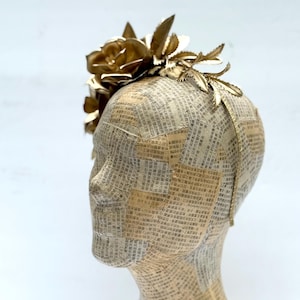 May include: A gold-colored floral headpiece with a large rose and leaf accents. The headpiece is attached to a mannequin head covered in newspaper. The headpiece is a decorative accessory.