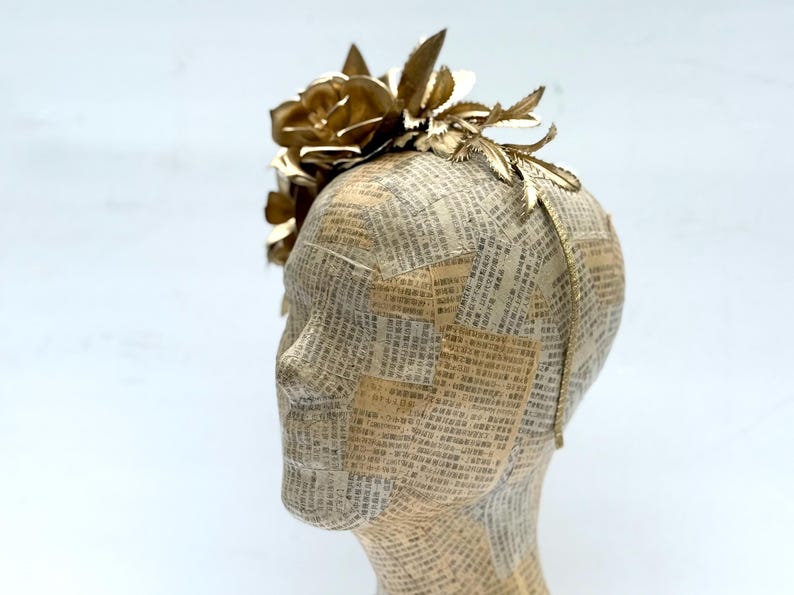 May include: A gold-colored floral headpiece with a large rose and leaf accents. The headpiece is attached to a mannequin head covered in newspaper. The headpiece is a decorative accessory.