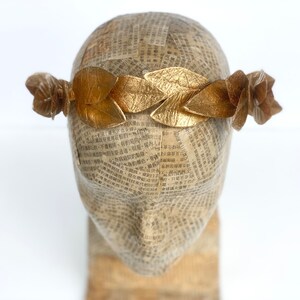 May include: A gold leaf crown with a simple design. The crown is made of individual leaves that are connected together. The crown is on a mannequin head.