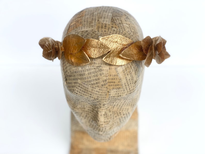 May include: A gold leaf crown with a simple design. The crown is made of individual leaves that are connected together. The crown is on a mannequin head.