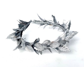 May include: A silver laurel wreath made of metal leaves. The wreath is a circle and is lying on a white surface.