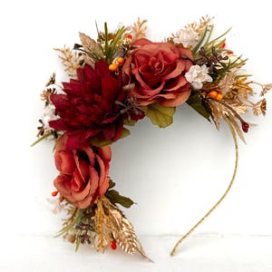 May include: A floral crown featuring burgundy and peach-colored roses, a dark red dahlia, and various greenery. The crown includes gold-colored accents and small orange berries, all set on a thin, gold-colored wire base.