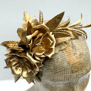 May include: A golden headpiece featuring large, metallic roses and leaves. The headpiece is adorned with intricate details and is set against a neutral background. The base is covered in newspaper print.