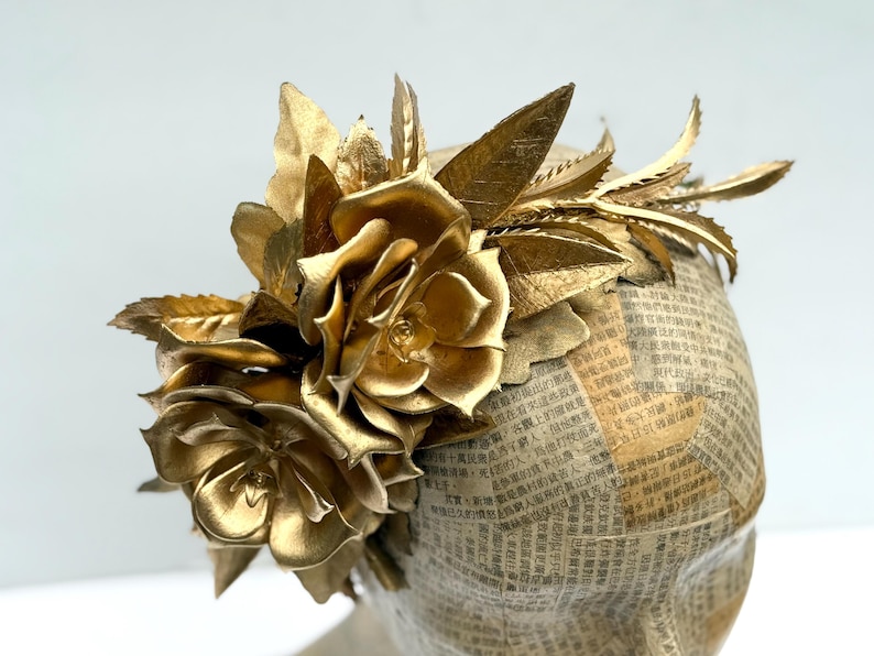 May include: A golden headpiece featuring large, metallic roses and leaves. The headpiece is adorned with intricate details and is set against a neutral background. The base is covered in newspaper print.