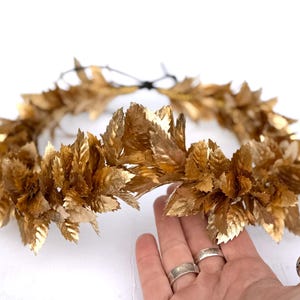 May include: A gold-colored leaf crown, likely for a special occasion. The crown is made of many gold-toned leaves, arranged in a circular shape. The leaves have a textured appearance, and the crown is set against a white background.