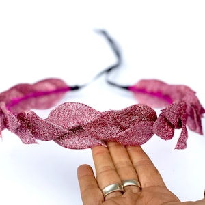 Könnte beinhalten: Ein festliches, glitzerndes, burgunderfarbenes Kopfstück mit einem geflochtenen Design. Das Kopfstück besteht aus einem schimmernden Material und hat einen schwarzen Krawattenverschluss. Das Design wird von einer Hand mit silbernen Ringen gehalten.