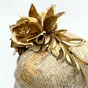 May include: A golden headpiece featuring a large, stylized flower and leaves. The floral elements are intricately designed and appear to be made of metallic material. The base is covered in newspaper print.
