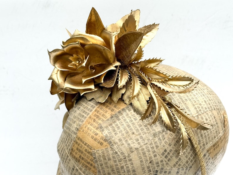 May include: A golden headpiece featuring a large, stylized flower and leaves. The floral elements are intricately designed and appear to be made of metallic material. The base is covered in newspaper print.