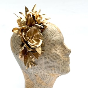 May include: A golden floral headpiece featuring large, stylized flowers and leaves. The headpiece is mounted on a mannequin head covered in newspaper print, creating a striking contrast. The flowers are metallic and reflect light.