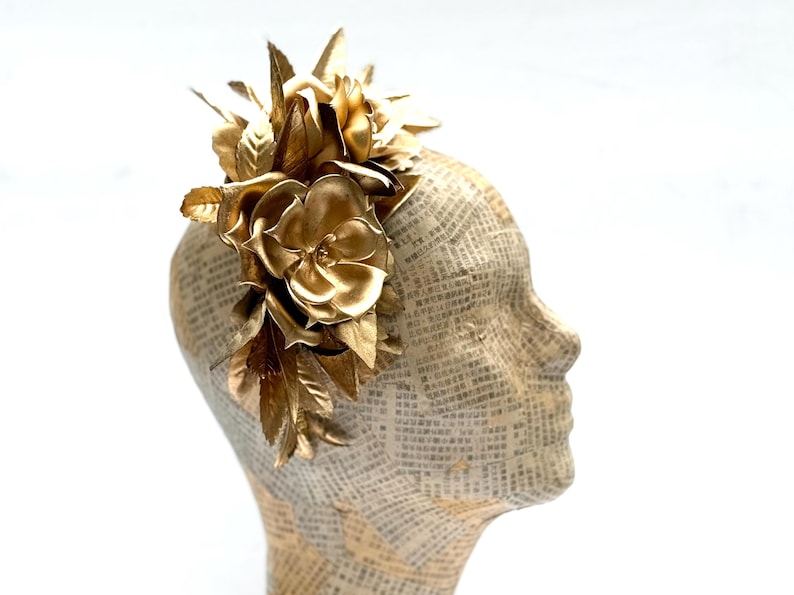 May include: A golden floral headpiece featuring large, stylized flowers and leaves. The headpiece is mounted on a mannequin head covered in newspaper print, creating a striking contrast. The flowers are metallic and reflect light.