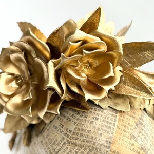 May include: A golden floral arrangement featuring two large, detailed roses and several leaves. The flowers are set atop a cylindrical base covered in newspaper print. The overall design is elegant and decorative.