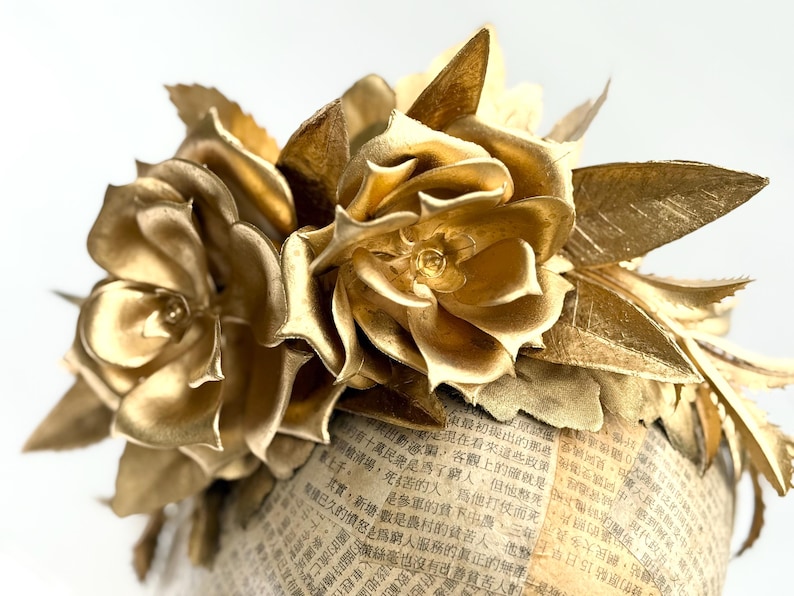 May include: A golden floral arrangement featuring two large, detailed roses and several leaves. The flowers are set atop a cylindrical base covered in newspaper print. The overall design is elegant and decorative.