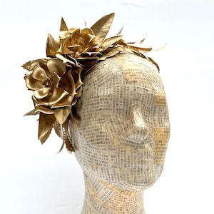 May include: A golden headpiece featuring large, stylized flowers and leaves. The floral arrangement is made of metallic gold material. The headpiece is displayed on a mannequin head covered in newspaper print.