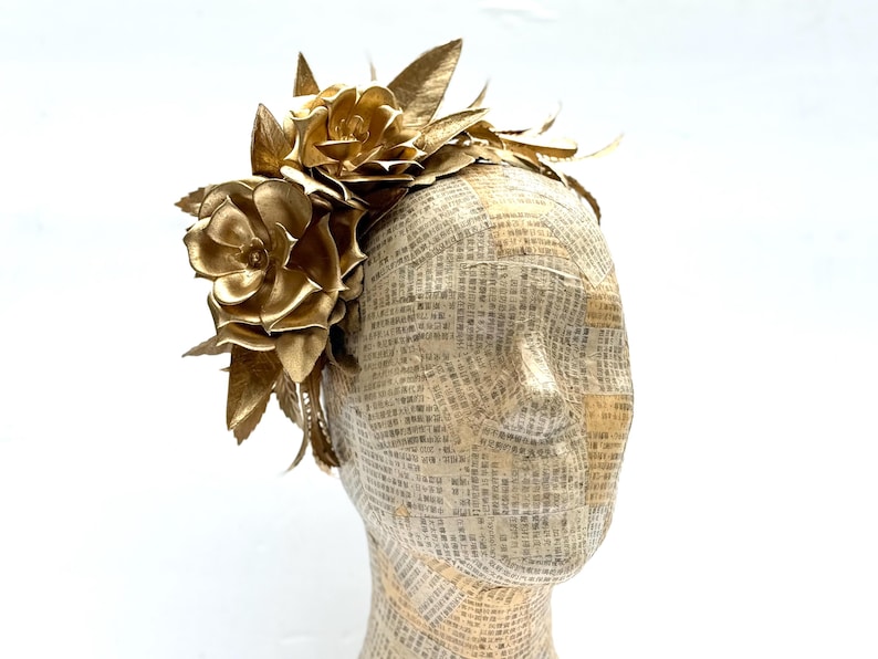 May include: A golden headpiece featuring large, stylized flowers and leaves. The floral arrangement is made of metallic gold material. The headpiece is displayed on a mannequin head covered in newspaper print.