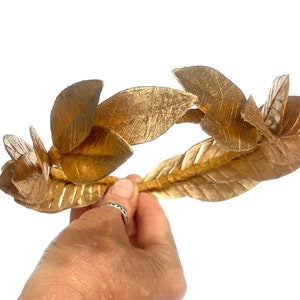 May include: A gold laurel wreath crown with leaves made of fabric. The crown is held in a hand.