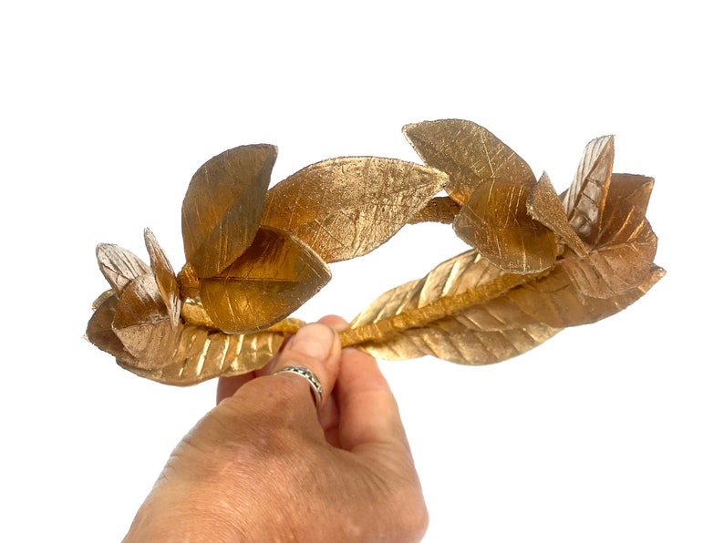 May include: A gold laurel wreath crown with leaves made of fabric. The crown is held in a hand.