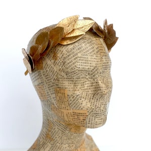May include: A gold leaf laurel wreath headband. The headband is made of individual gold leaves that are attached to a wire base. The headband is shown on a mannequin head.