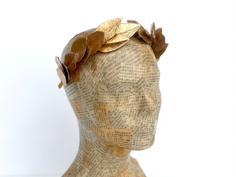 May include: A gold leaf laurel wreath headband. The headband is made of individual gold leaves that are attached to a wire base. The headband is shown on a mannequin head.