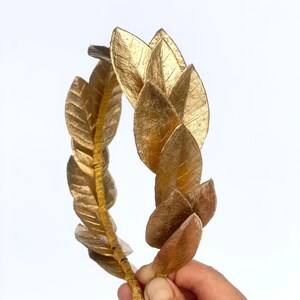 May include: A gold laurel wreath crown made of leaves. The crown is held in a hand.