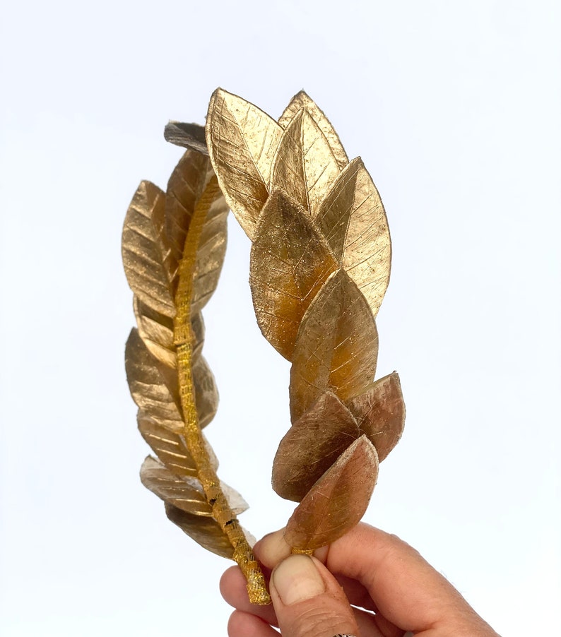 May include: A gold laurel wreath crown made of leaves. The crown is held in a hand.