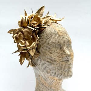 May include: A golden headpiece featuring large, detailed fabric flowers and leaves. The floral arrangement is attached to a head form covered in newspaper print. The headpiece is a striking accessory.