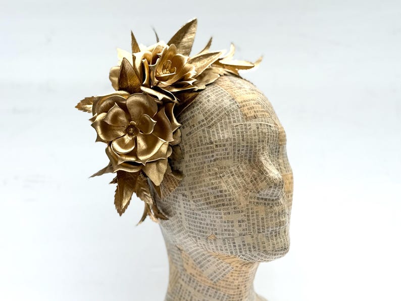 May include: A golden headpiece featuring large, detailed fabric flowers and leaves. The floral arrangement is attached to a head form covered in newspaper print. The headpiece is a striking accessory.