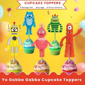Op de afbeelding: Een set kleurrijke Yo Gabba Gabba cupcake toppers. De toppers tonen personages zoals een blauw monster, een roze personage, een groen monster en een gele robot. Elke topper is geplaatst op een cupcake met kleurrijke sprinkles.