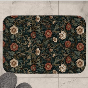 Può includere: Un tappetino da bagno rettangolare con un motivo floreale nei toni del verde, rosso e crema. Il tappetino è etichettato "SMALL" e misura 61 cm x 43 cm. Delle pantofole grigie sono posizionate vicino al tappetino su un pavimento piastrellato.