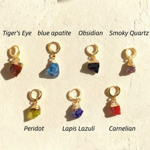 Puede incluir: Pendientes de aro dorados con colgantes de piedras preciosas variadas: Ojo de tigre, apatito azul, Obsidiana, Cuarzo ahumado, Peridoto, Lapislázuli y Cornalina. Cada pendiente presenta una piedra en bruto de diversos colores.