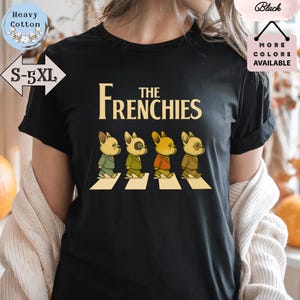 Peut inclure: T-shirt noir avec l'inscription "THE FRENCHIES" en or. Le motif représente quatre bouledogues français traversant un passage piéton, chacun portant une tenue de couleur différente. Le t-shirt est en coton épais et est disponible en tailles S-5XL.