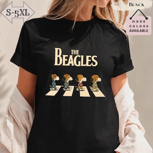 Peut inclure: Un t-shirt noir avec un motif représentant quatre chiens beagles marchant sur un passage piéton. Au-dessus des chiens, le texte THE BEAGLES est inscrit en lettres couleur crème. Les chiens portent des tenues variées en file indienne.
