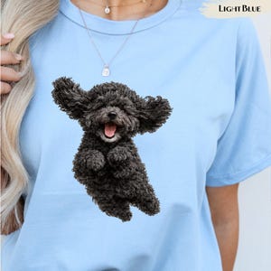 Op de afbeelding: Een lichtblauw t-shirt met een grafische print van een kleine, pluizige zwarte hond met wapperende oren en een open bek in een vrolijke uitdrukking. De tekst "LIGHT BLUE" staat in de rechterbovenhoek van het kledingstuk.